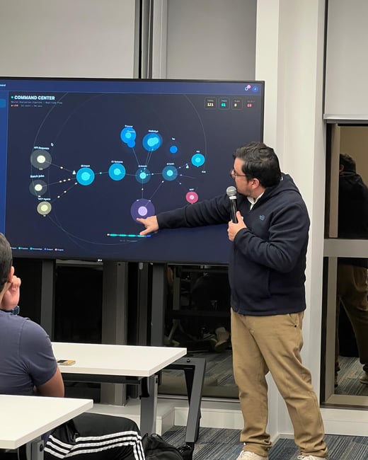 A man in a dark hoodie and khaki pants stands presenting in front of a large screen displaying a 'Command Center' network flow diagram, pointing at one of the nodes while holding a microphone.