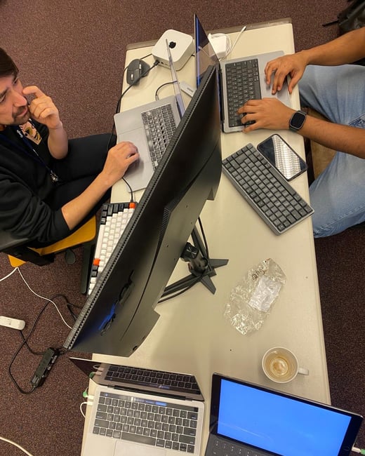 A top-down view of two individuals working at a desk crowded with various electronic devices, including multiple laptops, keyboards, a large monitor, and a Mac Mini.