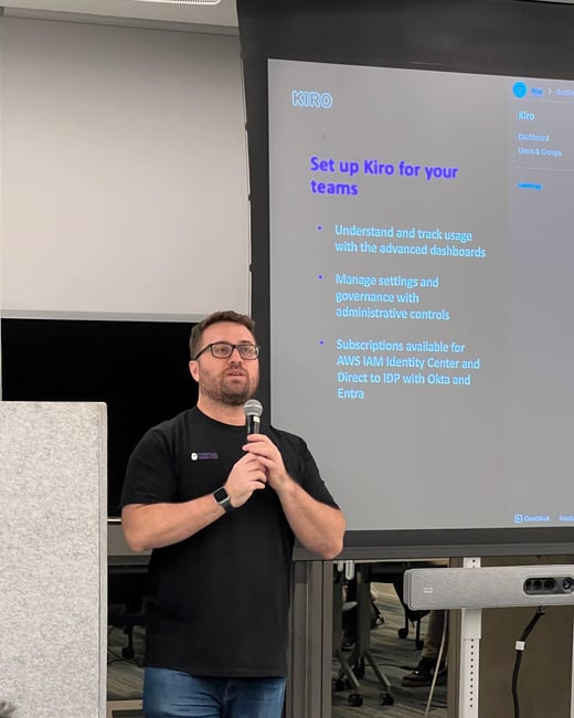 A man wearing glasses and a black t-shirt is presenting in front of a projection screen displaying a presentation about setting up Kiro for teams.