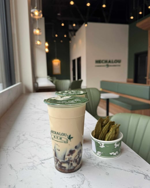 A close-up photograph of a plastic cup of milk tea (or similar dessert drink with jelly) and a small paper cup of green, leaf-shaped snacks, resting on a white, marble-patterned counter inside a modern cafe or tea house.