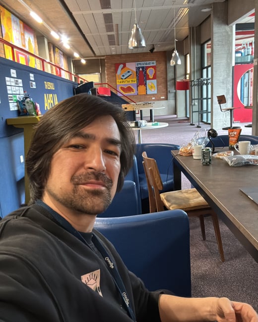 A man with medium-length brown hair and a goatee is taking a selfie while sitting at a table in a modern office or common area.