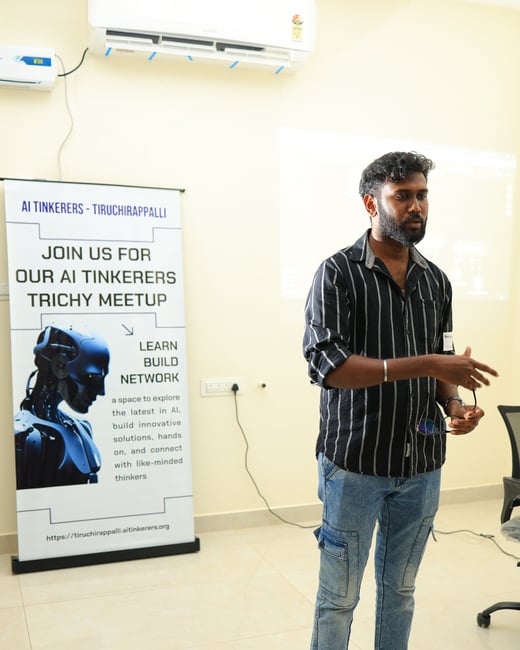 A man in a striped shirt and jeans stands barefoot next to an AI Tinkerers meetup banner in a room with an air conditioner.