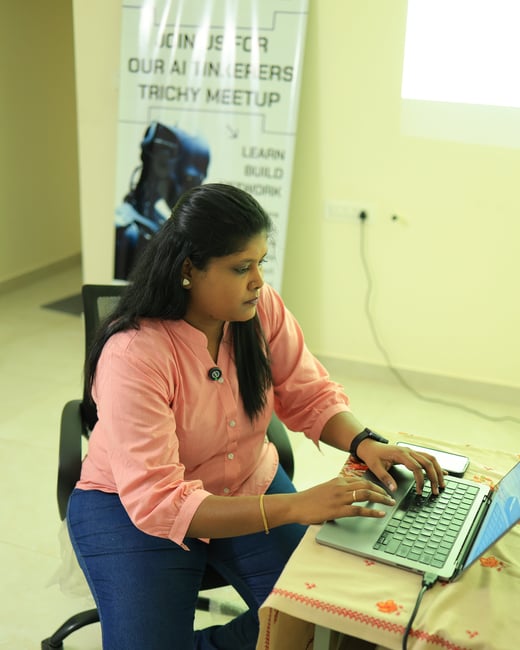 A woman in a pink shirt and blue jeans sits at a desk, focused on typing on a laptop. Behind her stands a promotional banner for an 'AI Engineers Trichy Meetup'.