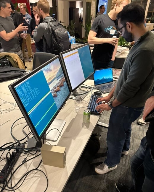 A candid photo capturing a tech event or hackathon setup, featuring a person working on a dual monitor display with code visible, while several other people mingle and socialize in the dimly lit background.
