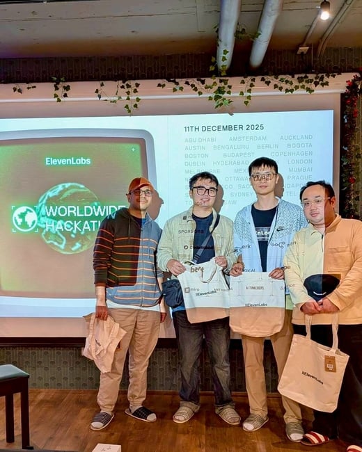 A photograph showing four men standing together in an indoor event space, posing in front of a projection screen displaying information about a worldwide hackathon organized by ElevenLabs and scheduled for December 11, 2025.