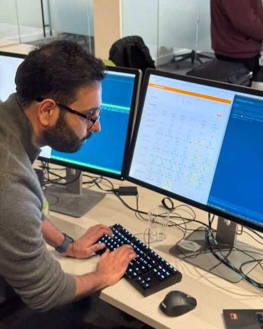 A man wearing glasses and a gray sweater is captured in profile, intently typing on a backlit mechanical keyboard while looking at dual computer monitors displaying technical data and dashboards in an office setting.