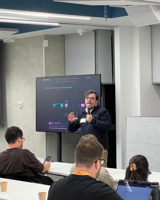 A man stands at the front of a modern presentation room, speaking into a microphone while gesturing towards a large screen displaying a diagram. An audience sits at white tables in the foreground, listening to the presentation.