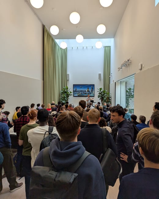 A vertical photograph showing a large crowd of people attending an indoor event or presentation in a modern, high-ceilinged room illuminated by spherical pendant lights.