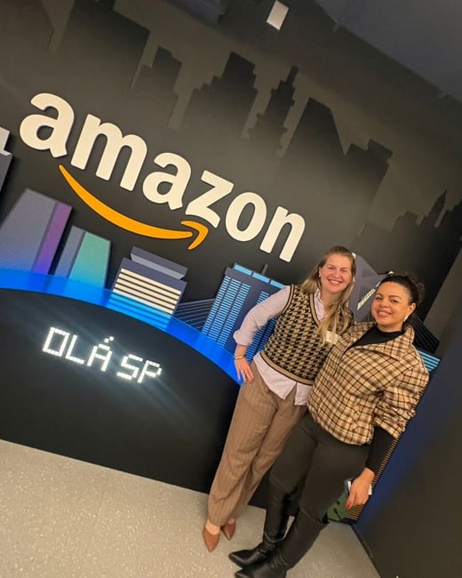Two women posing and smiling in front of an Amazon-themed backdrop with a city skyline graphic and the text 'OLÁ SP'.