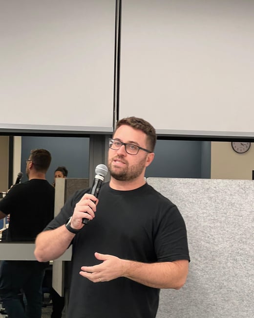A man wearing a black t-shirt and blue jeans is speaking into a wireless microphone during a presentation or meeting in an office setting.