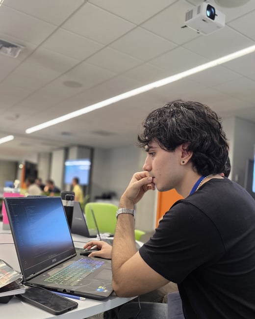 A young man with dark curly hair sits at a white desk, intently focused on a laptop screen displaying code in a modern office or event space.