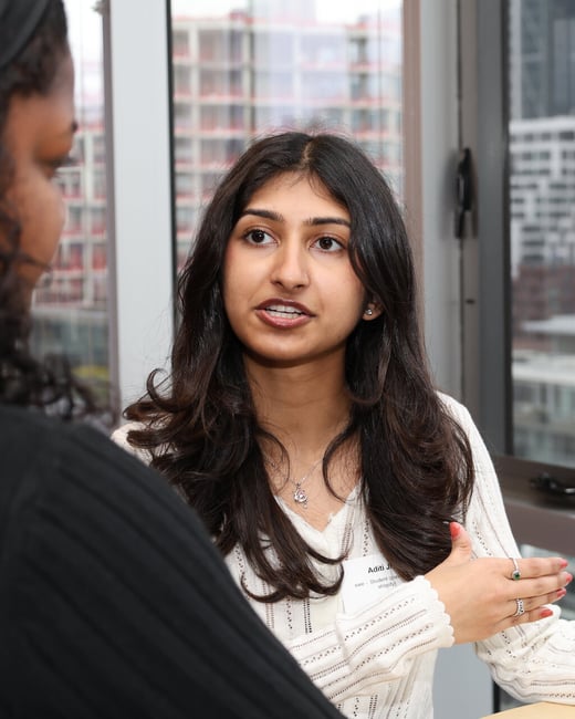 A young woman with long dark hair is engaged in a conversation, gesturing with her hand while wearing a name tag.