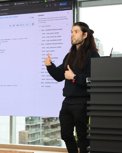 A man with long dark hair is giving a presentation in front of a large screen displaying software code in a modern office setting.