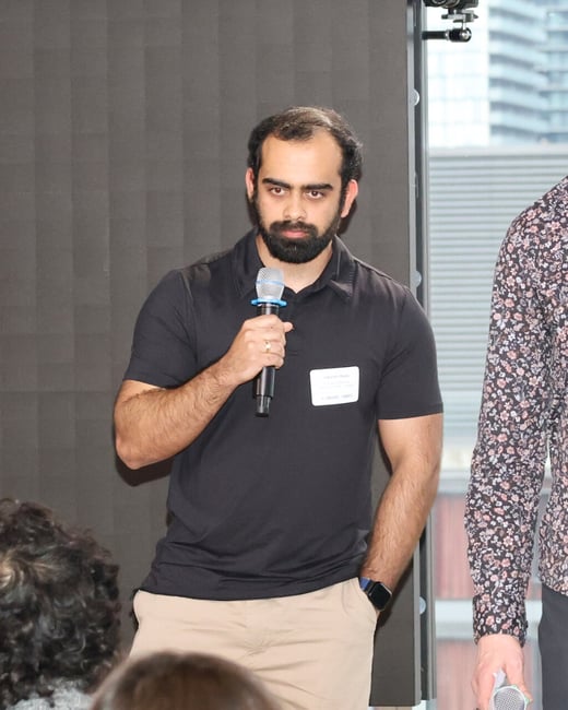 A man with a beard is speaking into a microphone at an event, wearing a black polo shirt and a name tag.