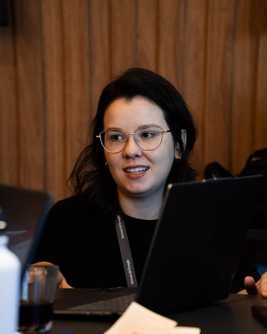A woman with glasses and dark hair is sitting at a table with a laptop, wearing a 'Women@Google' lanyard.