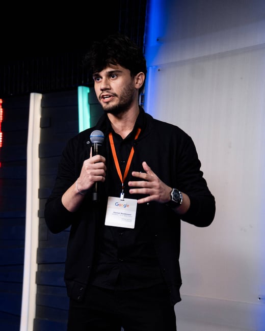 A young man with dark hair and a beard is speaking into a microphone while gesturing with his hand during a presentation at a Google event.