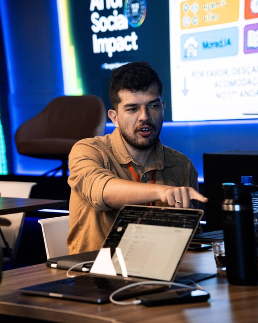 A man in a tan shirt sits at a table with a laptop, pointing at something while speaking during an event with a screen displaying 'AI for Social Impact' in the background.
