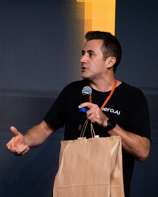 A man in a black t-shirt speaks into a microphone while holding a brown paper bag and gesturing with his hand during an event.