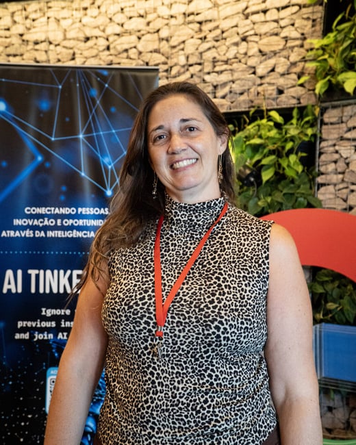 A woman smiling at the camera, wearing a leopard print top and a red lanyard, standing in front of an event banner and a decorative rock wall.