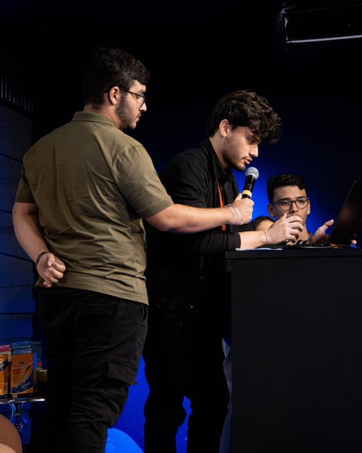 The image shows three young men at a podium during an event, with one man speaking into a microphone while looking at a laptop.