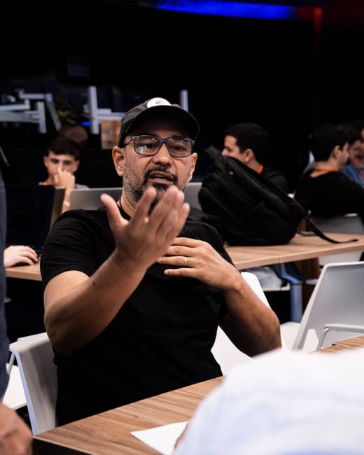 A man wearing glasses and a baseball cap is gesturing with his hand while engaged in a conversation at a table in an indoor setting.