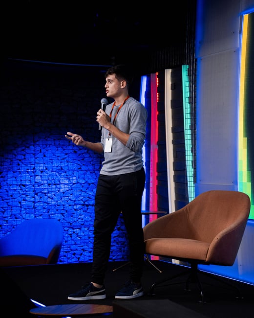 A young man stands on a stage with a microphone, gesturing while giving a presentation in a modern studio setting with colorful lighting.