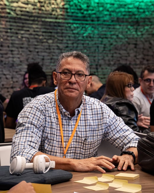 A man in a blue and white checkered shirt and glasses sits at a table with sticky notes and headphones in a crowded indoor setting.