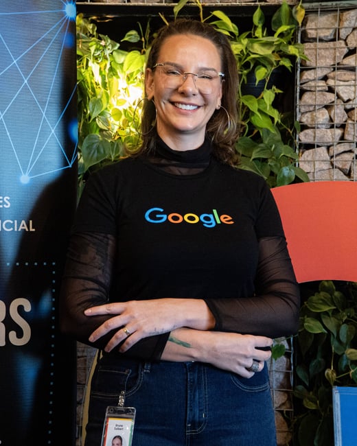 A woman in a black Google t-shirt and glasses smiles for a portrait in front of a wall of plants and stones.