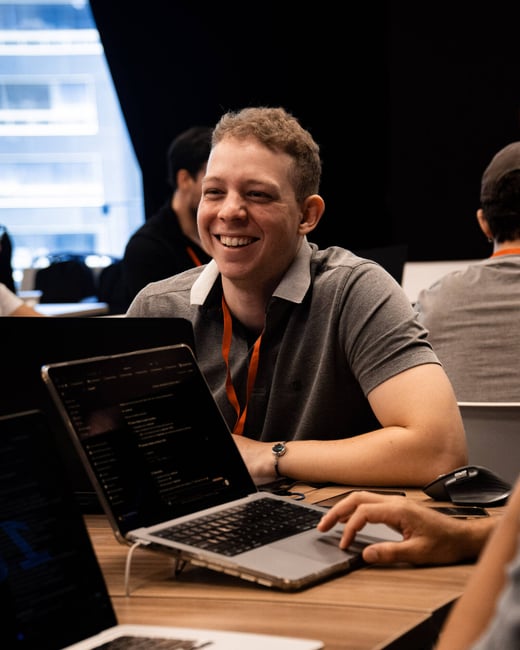 A young man with short curly hair is smiling warmly while sitting at a wooden table with several open laptops, likely during a tech event or workshop.