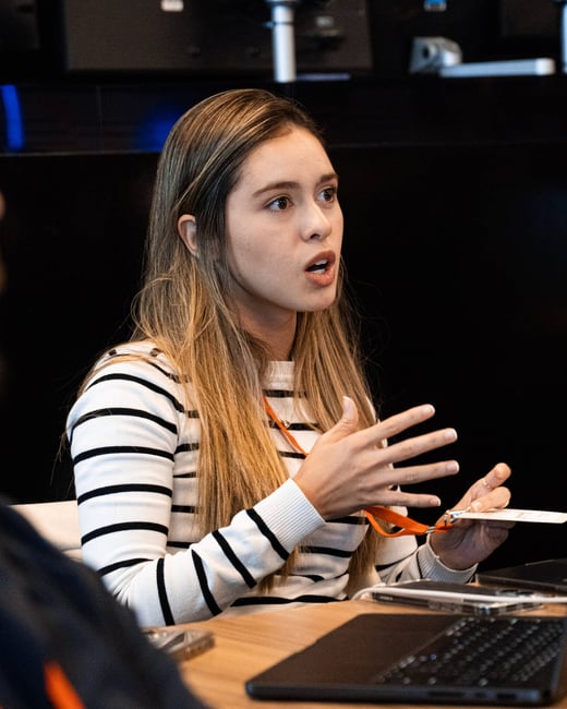 A young woman with long hair wearing a striped sweater is gesturing with her hands while speaking in an indoor setting.