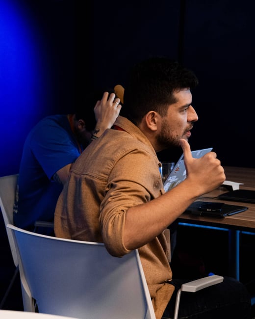 A man in a brown shirt is seated at a table, giving a thumbs-up gesture while looking towards someone off-camera.
