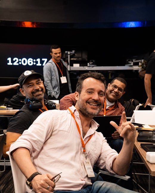 A group of men are smiling and posing for a photo in an indoor event or office space, with one man in the foreground making a hand gesture.