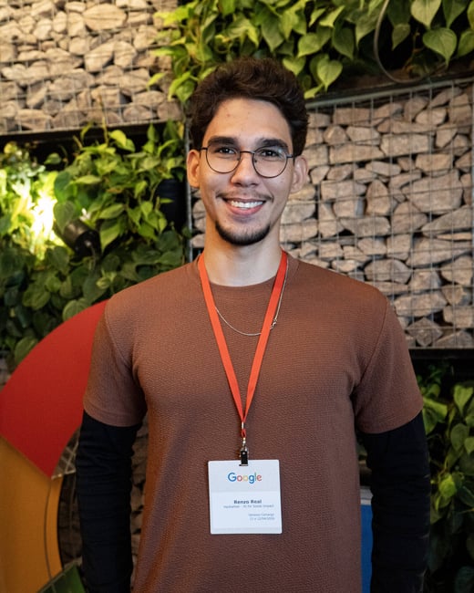 A portrait of a smiling young man wearing glasses and a brown t-shirt, standing in front of a wall decorated with plants and stones.