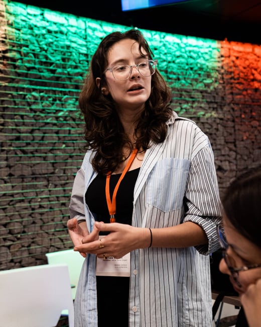 A woman wearing glasses and a striped shirt is speaking and gesturing at an event, standing in front of a stone wall illuminated with green and red lights.