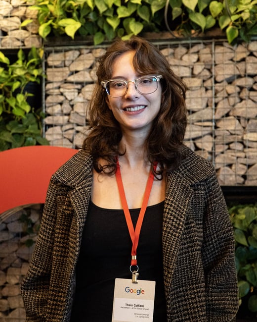A portrait of a smiling woman with glasses and wavy brown hair, wearing a Google event lanyard and a patterned coat in front of a stone and plant wall.
