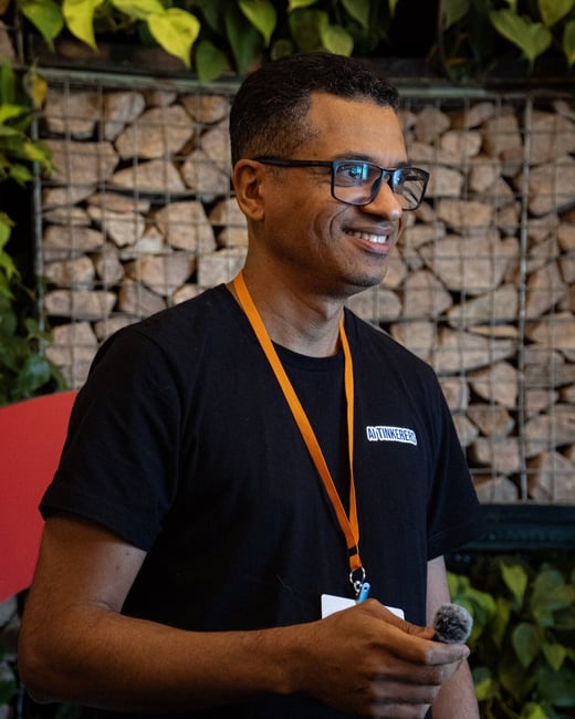 A smiling man wearing glasses and a black t-shirt with 'AI TINKERERS' printed on it stands in front of a stone wall background, holding a small microphone.