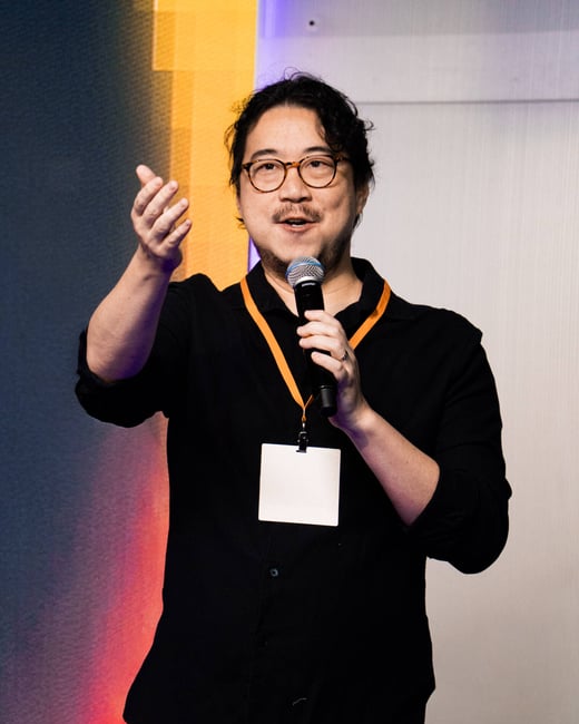 A man with glasses and a mustache is speaking into a microphone and gesturing with his hand during an indoor event.