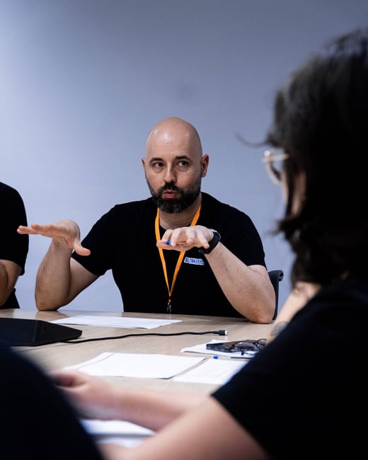 A man with a beard and bald head gestures with his hands while sitting at a table during a meeting or discussion.