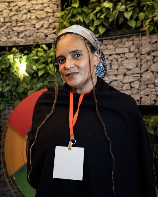 A portrait of a woman wearing a black top and a patterned headscarf, standing in front of a decorative wall featuring stones and green plants.