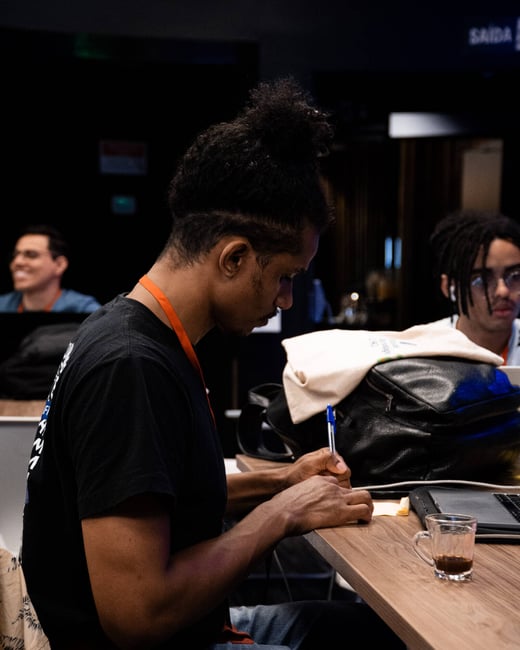 A young man with a bun hairstyle is sitting at a wooden table, focused on writing with a pen. In the background, other people are visible in a dimly lit indoor setting.