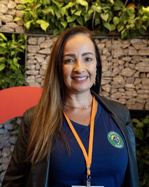 A portrait of a smiling woman with long brown hair wearing a blue t-shirt, a black jacket, and an orange lanyard, standing in front of a stone and plant wall.