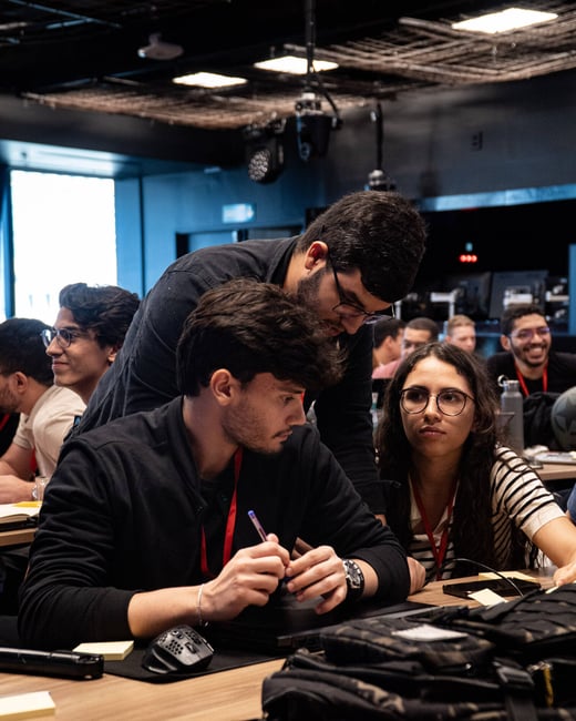 A group of young adults are focused on a task at a table during a tech event or workshop, with one man leaning over to assist others.