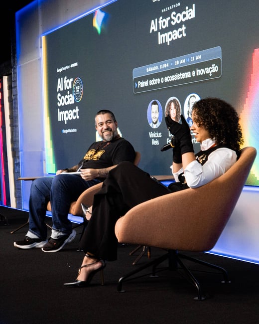 A man and a woman are seated on stage during a panel discussion at an 'AI for Social Impact' hackathon event.