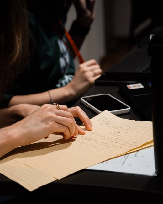A close-up photograph of hands resting on a desk next to a yellow legal pad with handwritten notes in Portuguese.