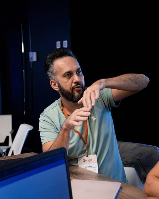 A man with a beard and dark hair is sitting at a table, gesturing expressively with his hands while wearing a Google event badge.