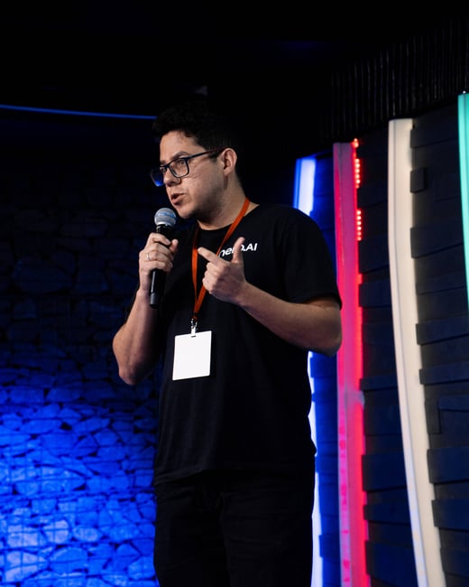A man wearing glasses and a black t-shirt is speaking into a microphone on a stage with vibrant blue and red lighting.