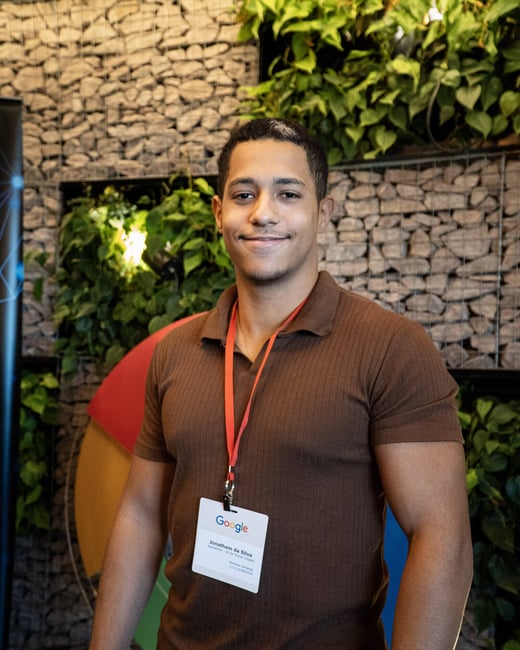 A portrait of a young man smiling, wearing a brown polo shirt and a Google event identification badge around his neck.