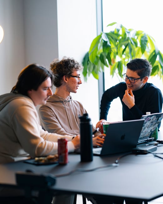 Three young men are gathered around a table with laptops, engaged in a collaborative discussion in an office setting with a large window and a plant in the background.