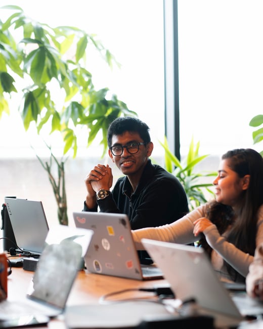 A photograph of a young man with glasses smiling at the camera while sitting at a wooden table with laptops, alongside a young woman looking to the side, set against a bright window with green plants.