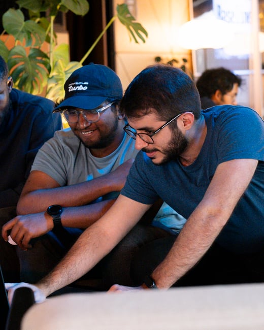 Two men are sitting close together, looking intently at a screen off-camera. One man wears a black Porsche Turbo cap and glasses while smiling, and the other wears a blue t-shirt and glasses as he leans forward.